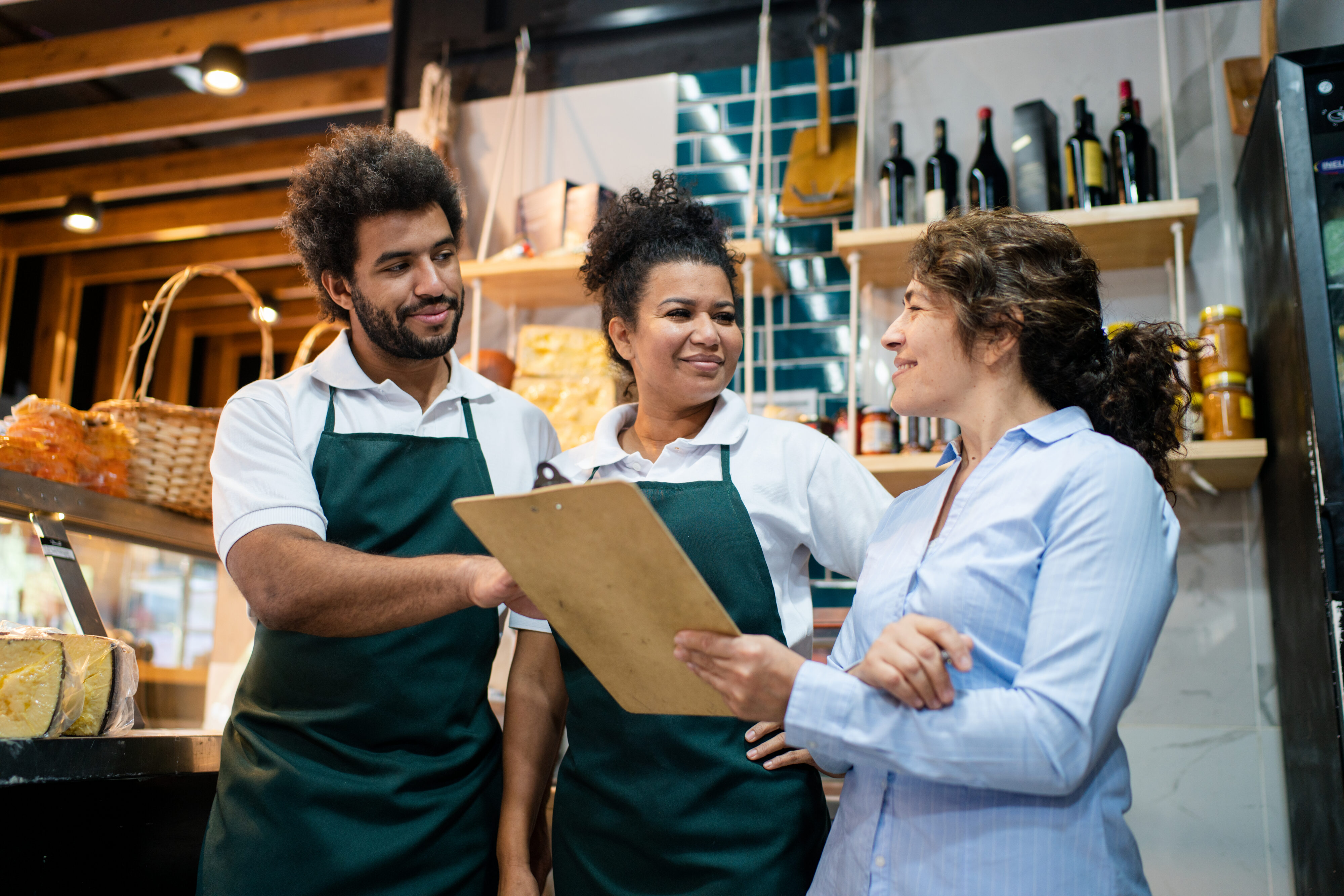 Female grocery store manager holding a checklist, discussing stock and products with male and female workers in the delicatessen section, ensuring inventory accuracy and quality.